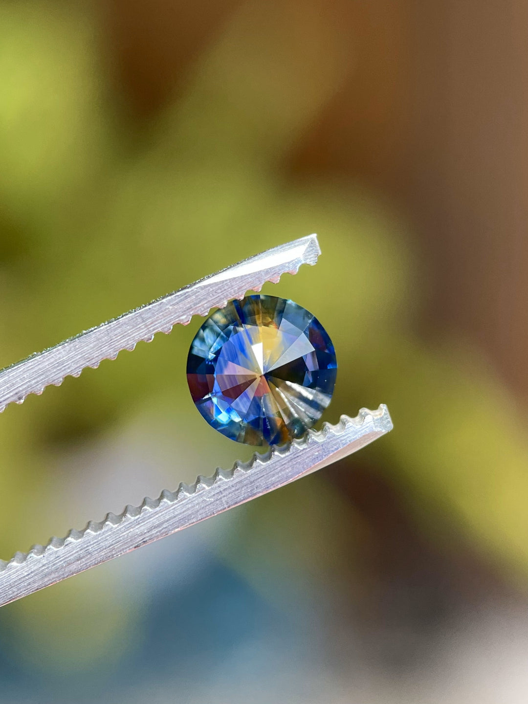 Close-up view of a 1.11-carat round brilliant parti sapphire held by tweezers, showing its distinct blue and yellow zoning under magnification.