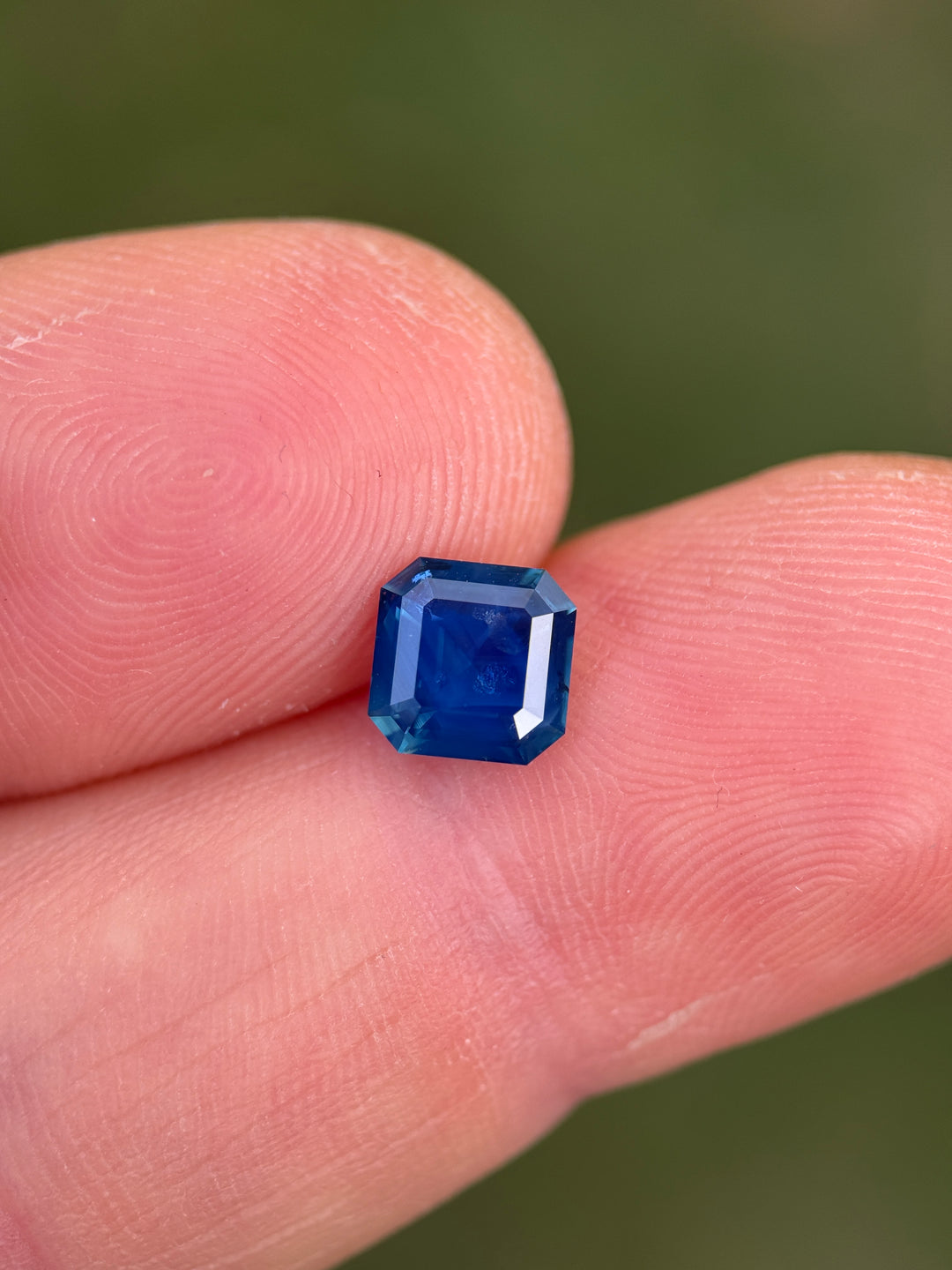 A small square-cut deep blue gemstone held between two human fingers against a blurred green background.