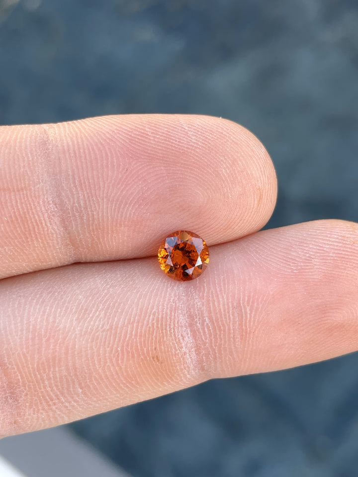 Vivid orange-brown Hessonite Garnet, 1.25ct, displayed between fingers for scale with a stunning round brilliant cut.