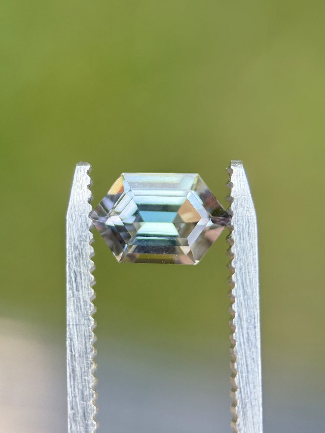 Mermaid-color Tanzanite gemstone held in tweezers outdoors, displaying its custom hexagon step cut and untreated clarity from Merelani Hills
