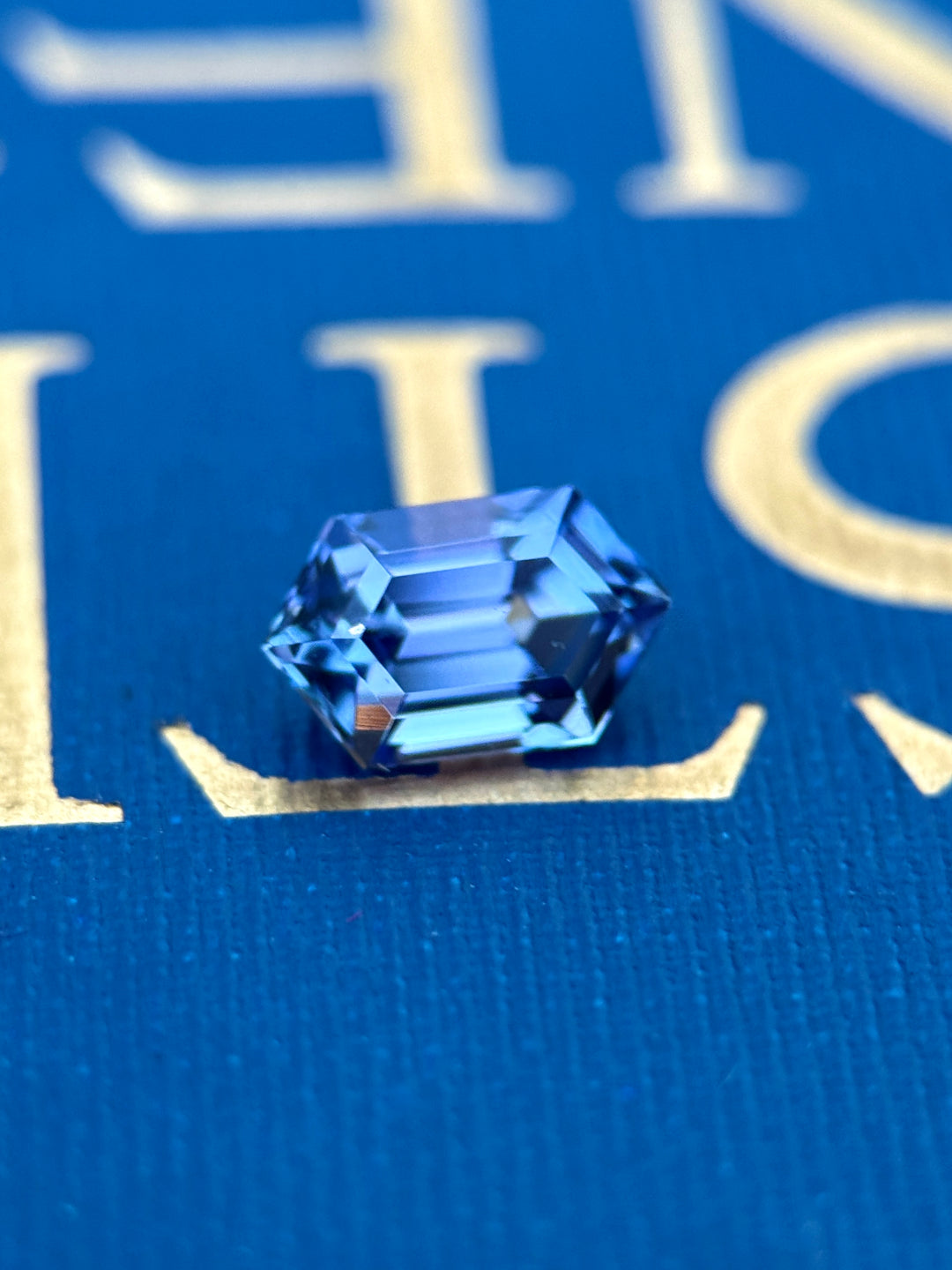 Untreated 1.43-carat hexagon step cut tanzanite with mermaid colors—blue, green, pink, and purple—resting on a blue and gold book cover, showcasing its reflective facets and multicolor shifts from Merelani Hills, Tanzania.