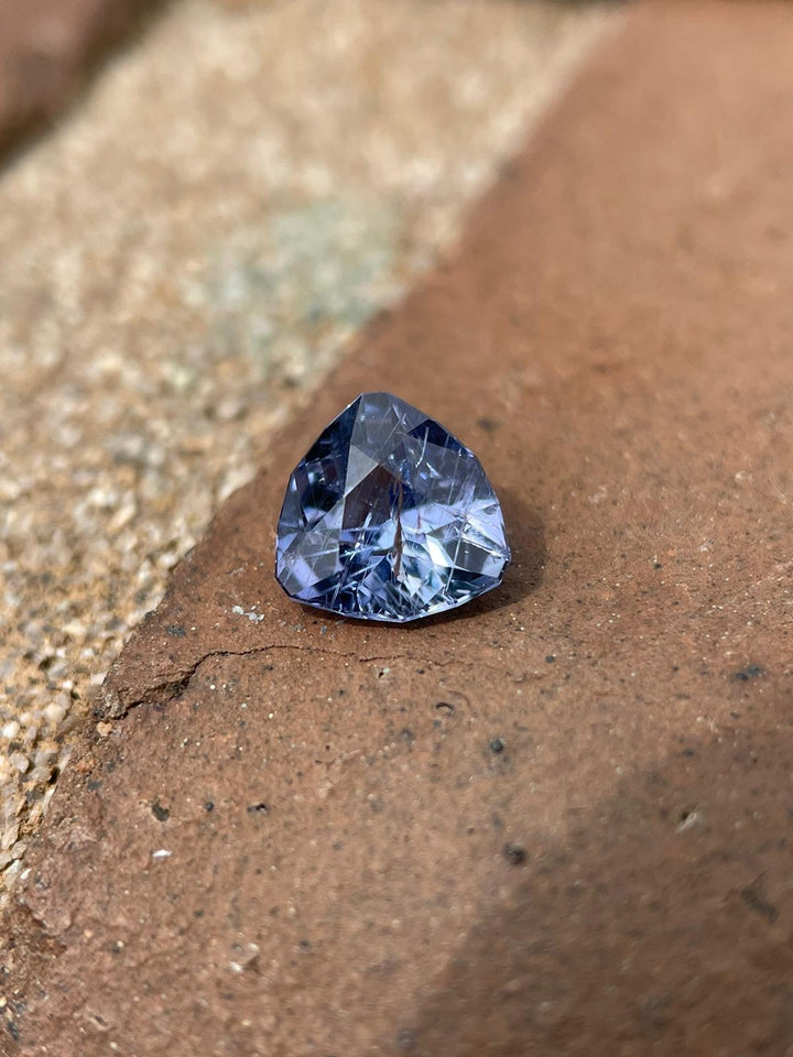 A stunning 1.38 carat custom trillion-cut sapphire, with vibrant violet and denim hues, resting on a textured stone surface, highlighting its exquisite facets and depth of color.