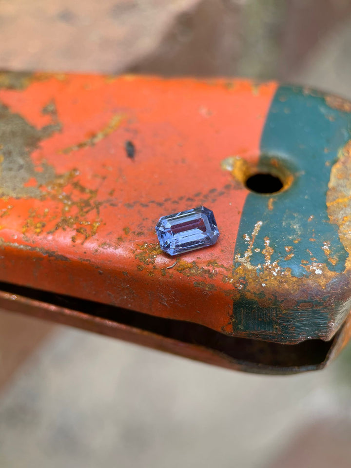 Violet-blue sapphire resting on an orange rusty surface, highlighting the gem's clarity and modified emerald cut, emphasizing its natural origin from the Umba River, Tanzania.