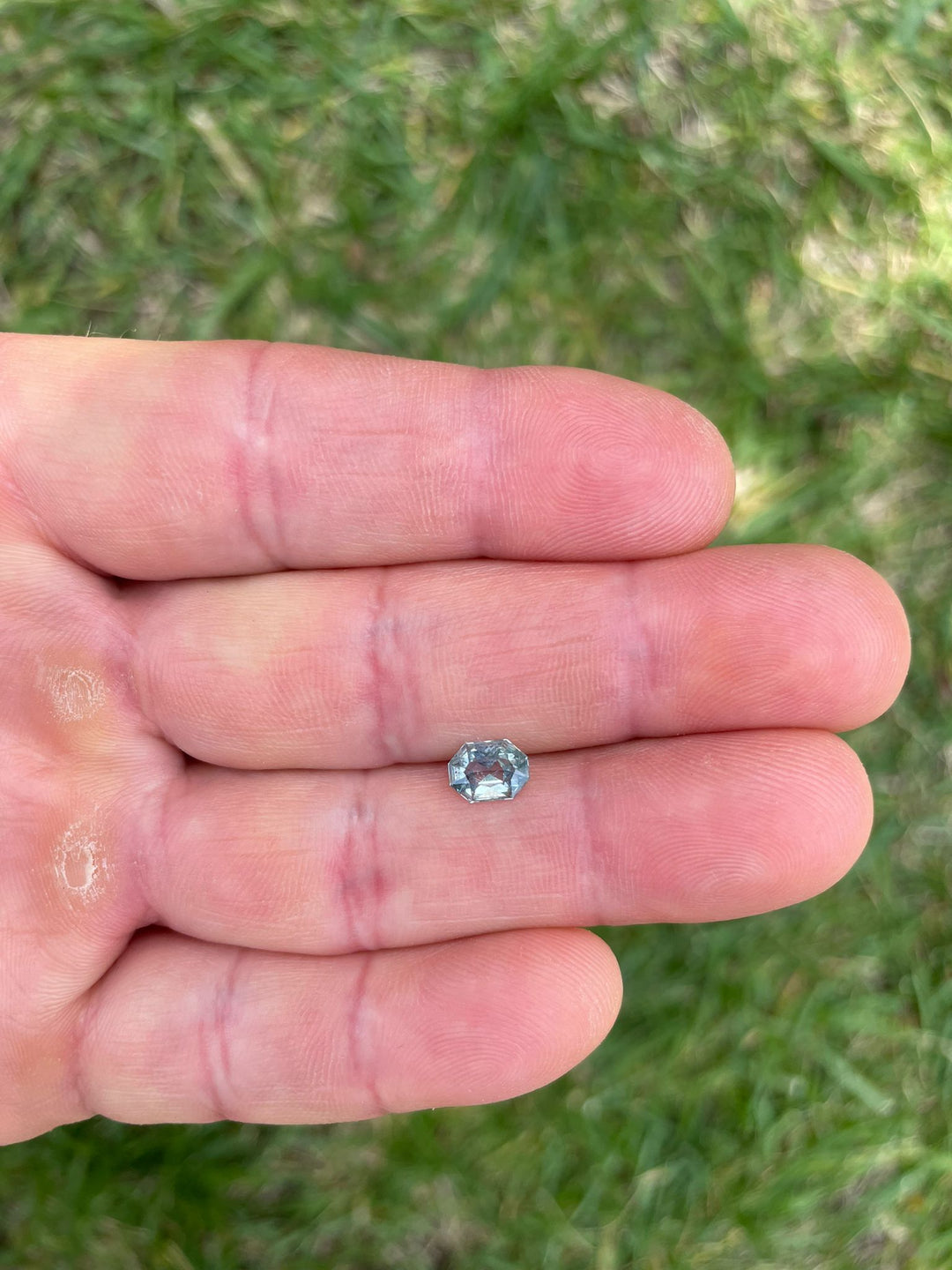 Hand holding a 0.86 carat yellow and seafoam sapphire with custom octagonal radiant shape from Tanzania, presented over a grassy background.