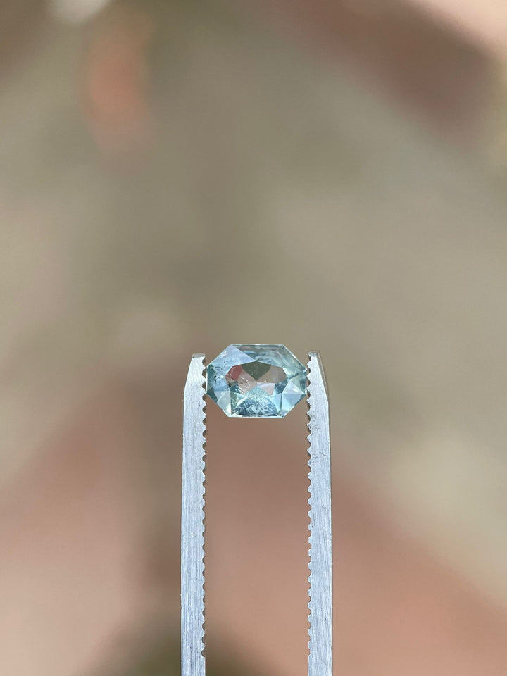 0.86 carat Tanzanian yellow and seafoam sapphire with custom octagonal radiant cut, displayed securely in a tweezer grip against a blurred background.