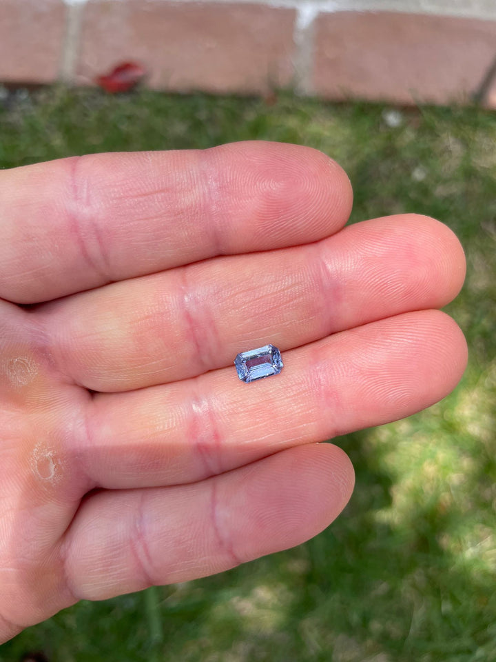 A violet-blue sapphire gemstone of 1.26 carats held between fingers, showcasing its modified emerald cut and vibrant hues against a natural background.