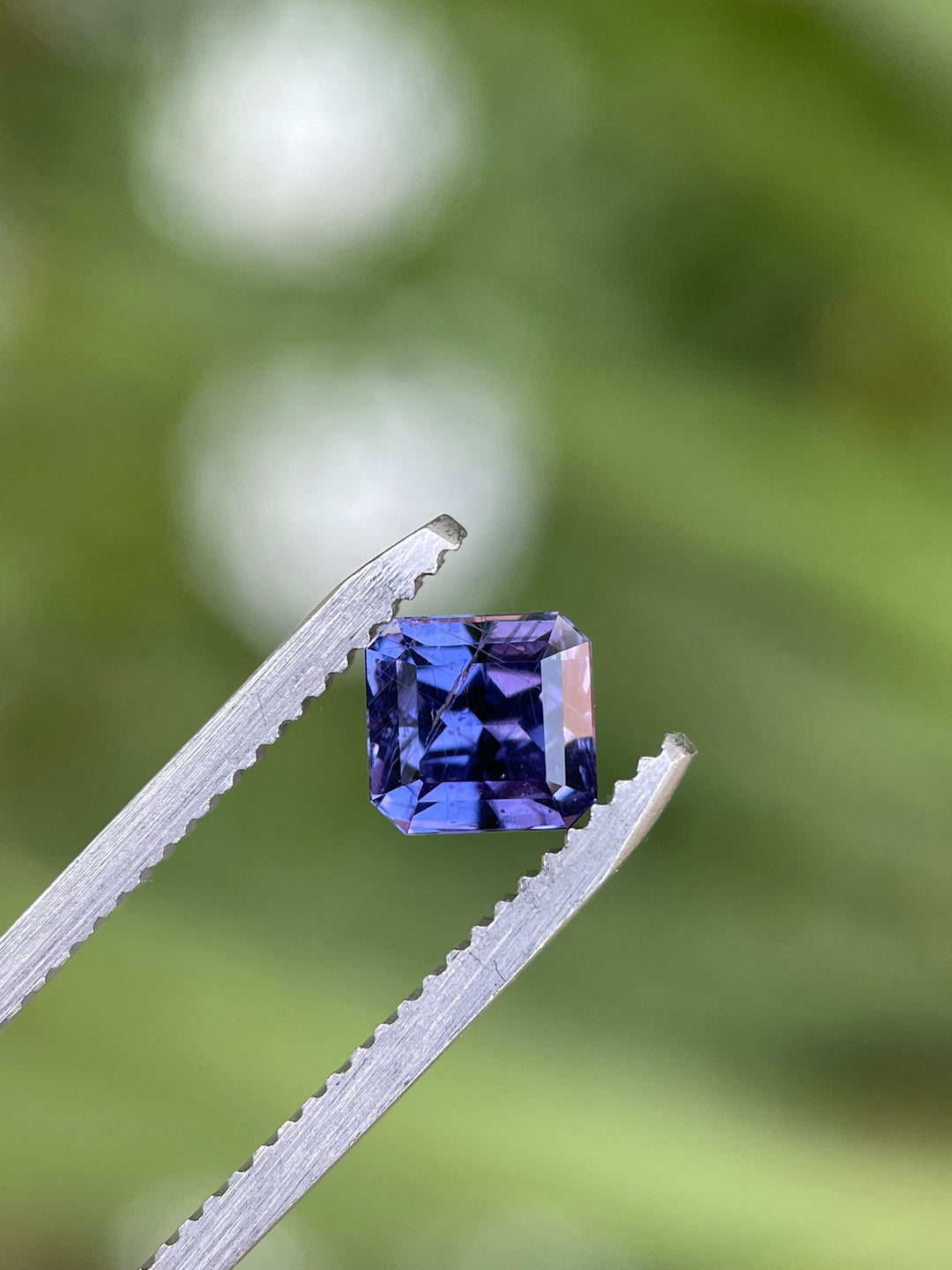Close-up view of a custom octagonal step cut sapphire held in tweezers, with a focus on its deep violet and purple hues against a blurred green background.