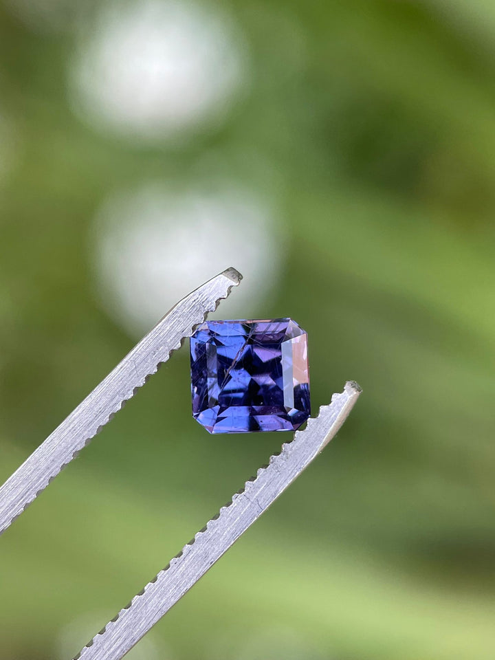 Close-up view of a custom octagonal step cut sapphire held in tweezers, with a focus on its deep violet and purple hues against a blurred green background.