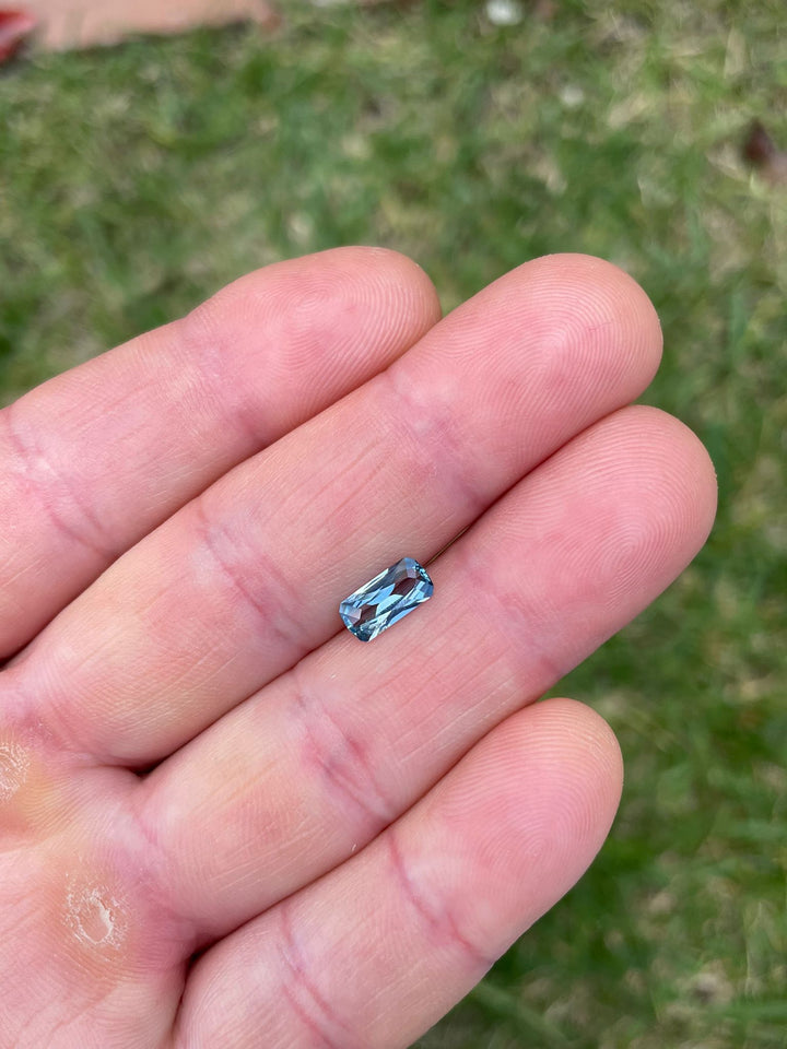 A vibrant blue sapphire rests on a human palm, showcasing its unique custom elongated geometric cushion cut. The gem weighs 1.26 carats and measures 8.97 x 4.50 x 2.99 mm, hailing from the Umba River in Tanzania. It is untreated, highlighting its natural beauty, valued at $1197.