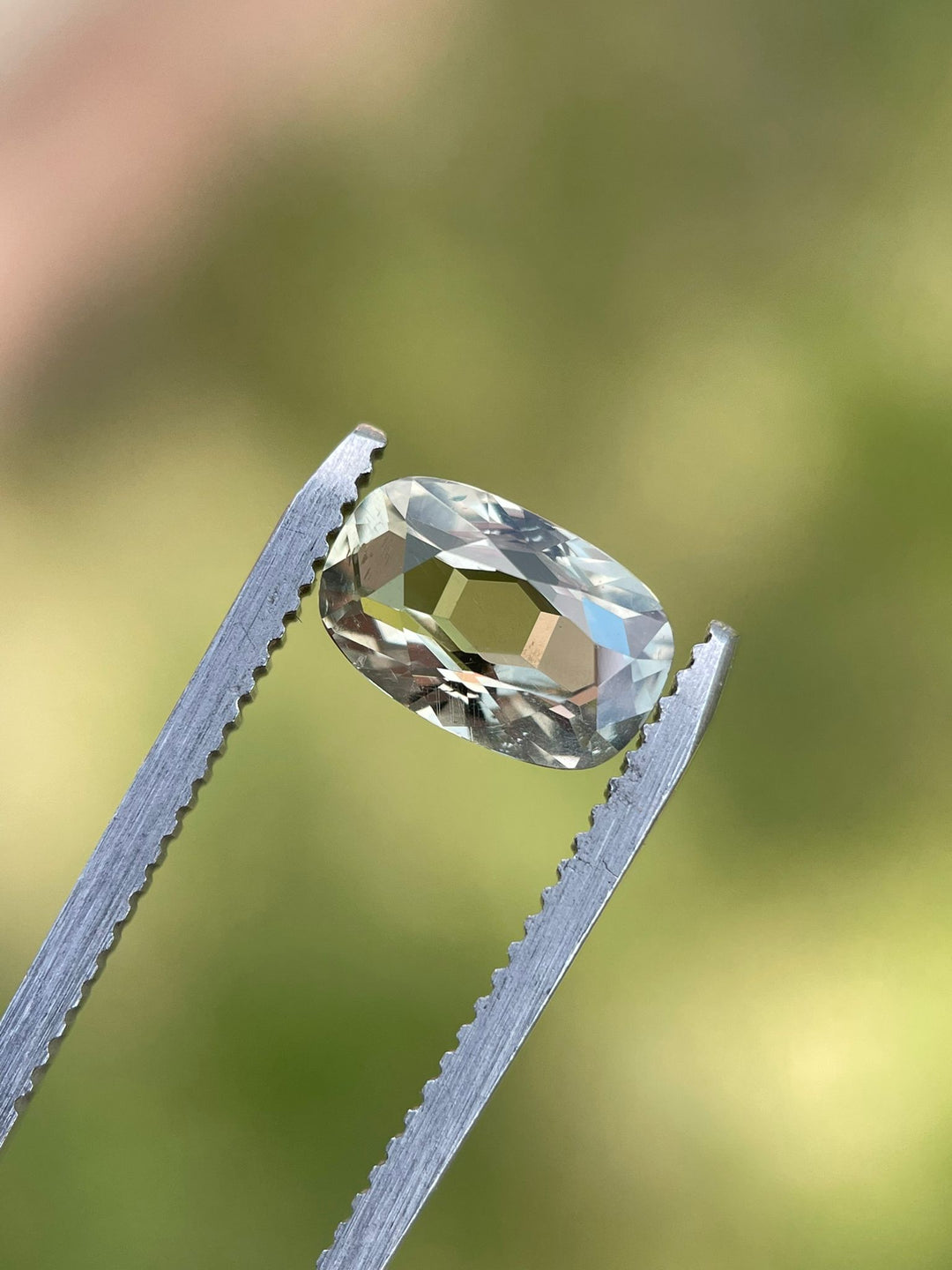 2.29-carat sapphire in a modified cushion cut, suspended in tweezers against a blurred green background, highlighting its striking color transitions from yellow to teal and green, untreated and Tanzanian origin.