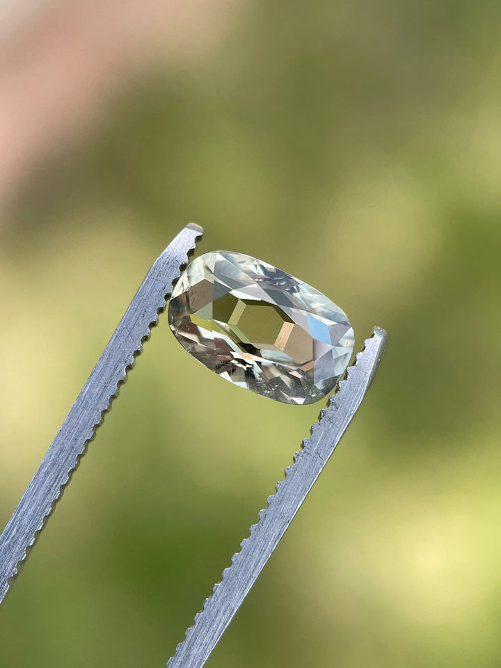 2.29-carat sapphire in a modified cushion cut, suspended in tweezers against a blurred green background, highlighting its striking color transitions from yellow to teal and green, untreated and Tanzanian origin.