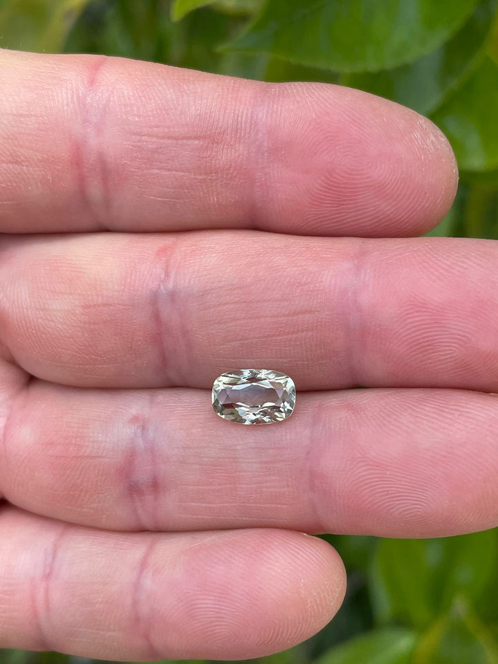 Close-up view of a 2.29-carat modified cushion cut sapphire, held between fingers, displaying a blend of yellow, teal, and green colors, with a detailed view of its facets, from Tanzania, natural.