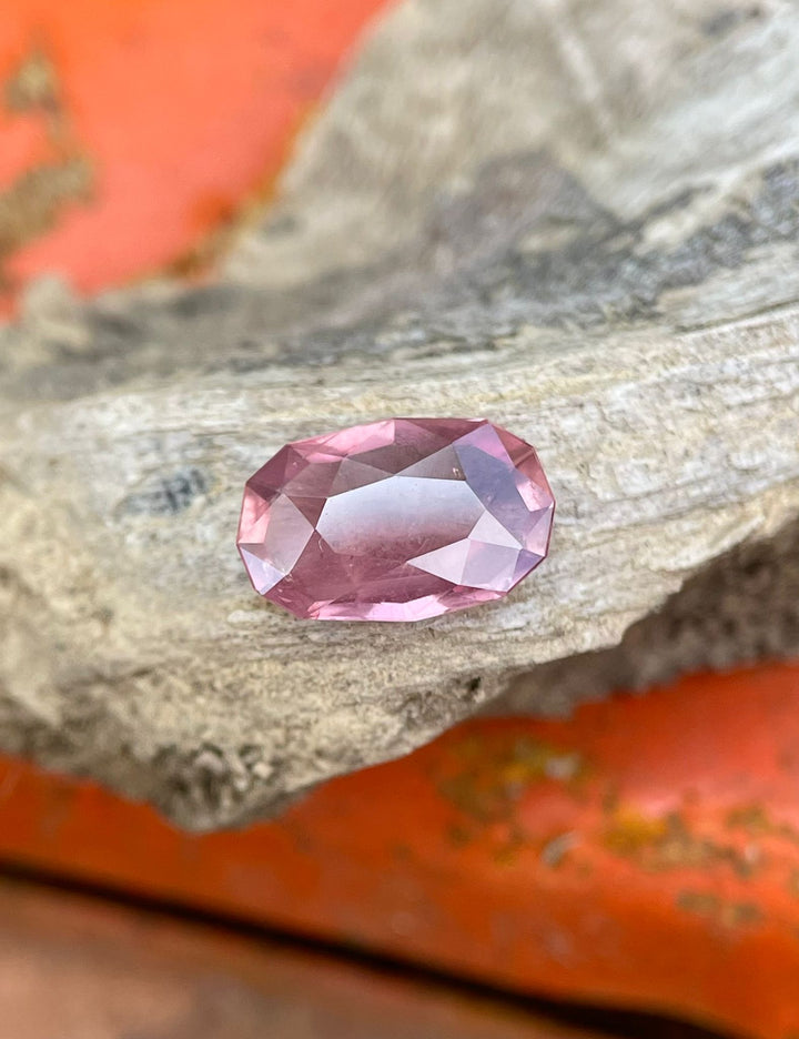 1.45 carat custom elongated cushion cut sapphire, displaying a unique grapefruit and pastel hue, held gently in a hand against a natural green background.