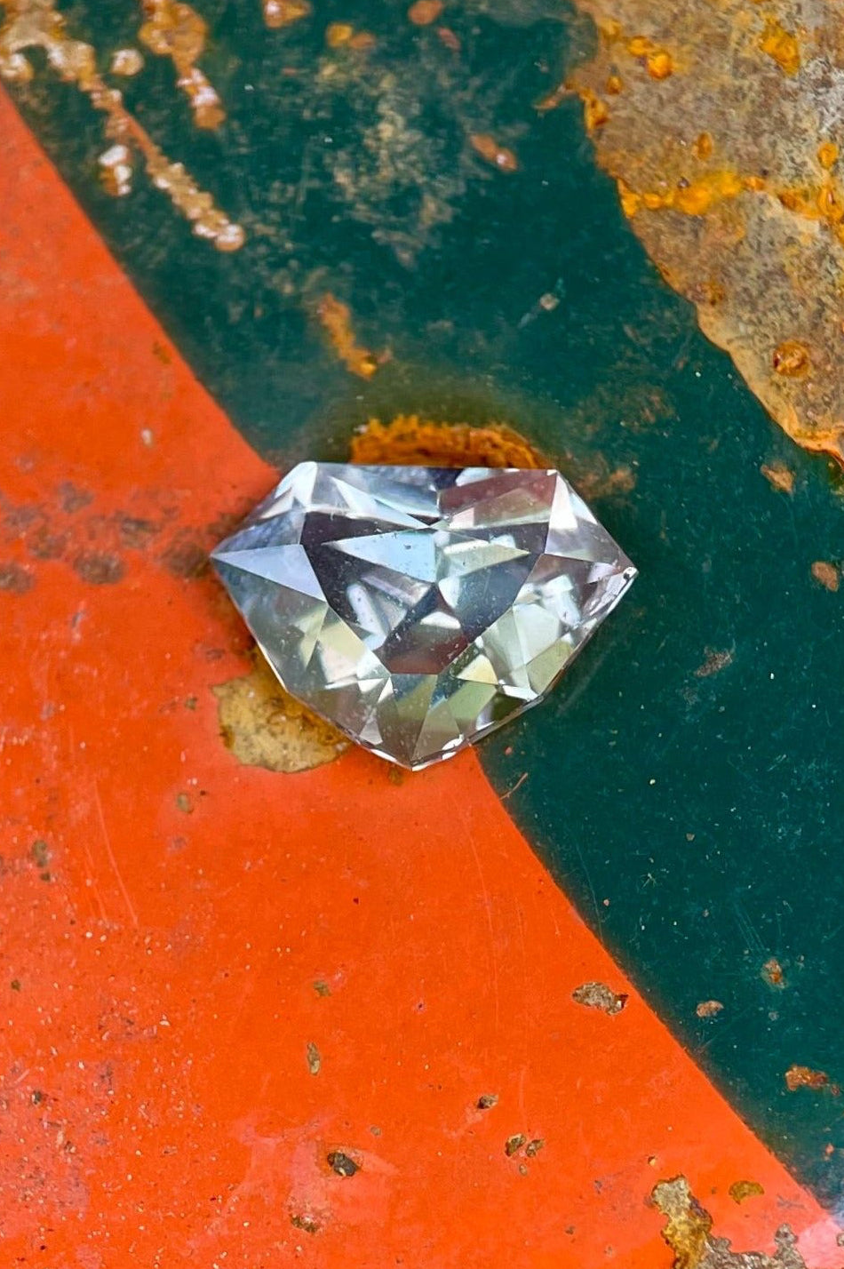 2.11 carat sapphire with a custom shield cut, showcasing a unique blend of gray, yellow, and teal colors, placed on a rusty and colorful surface, emphasizing its geometric beauty.