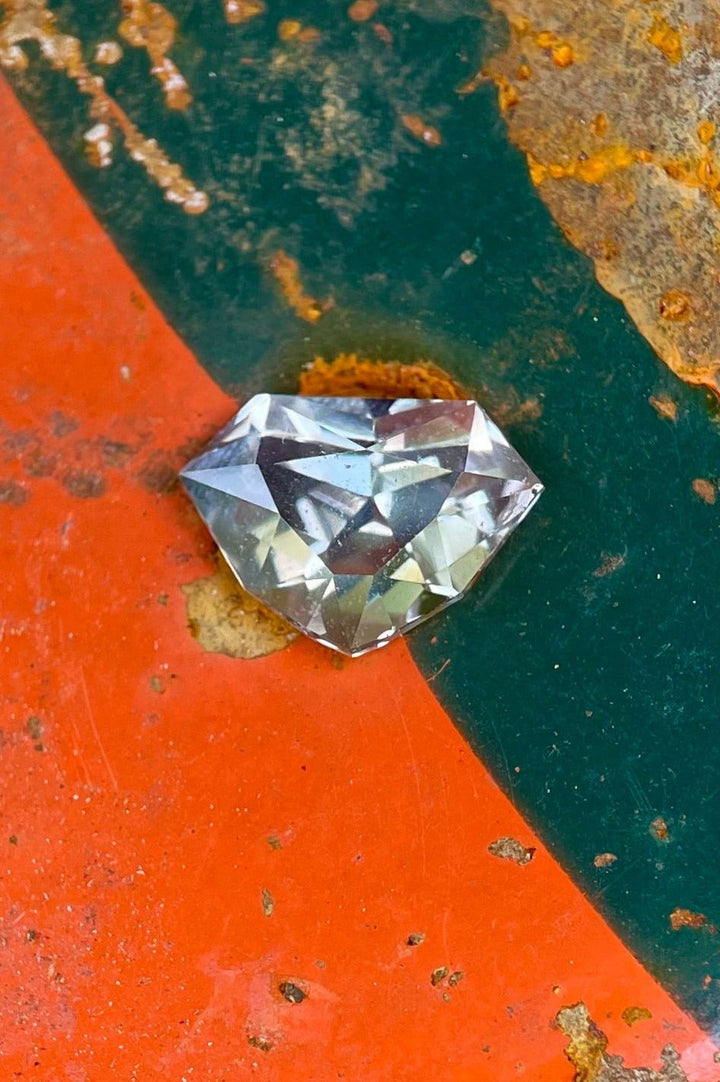 2.11 carat sapphire with a custom shield cut, showcasing a unique blend of gray, yellow, and teal colors, placed on a rusty and colorful surface, emphasizing its geometric beauty.