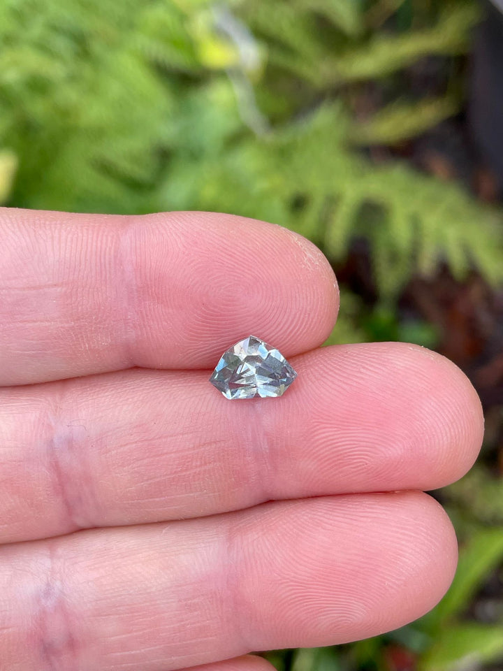 Close-up of a multicolored sapphire, featuring gray, yellow, and teal hues, held in fingers against a lush green background, highlighting its distinct shield shape.