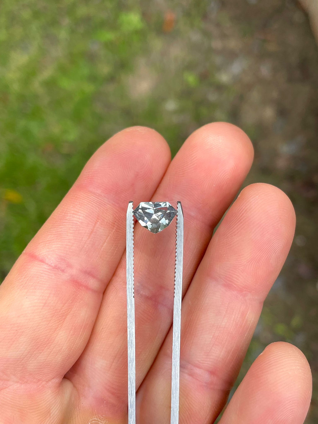 2.11 carat sapphire with a custom shield cut, delicately held in tweezers, displaying its striking gray, yellow, and teal colors against a natural backdrop." Title: "Custom Shield Cut Sapphire in Tweezers