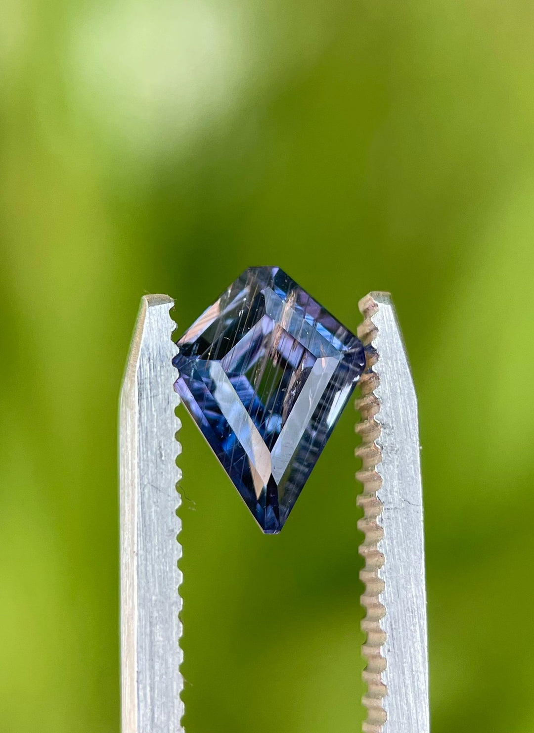 Custom step cut kite portrait sapphire, displaying a mesmerizing blend of teal, purple, and blue, positioned in tweezers with a soft-focus greenery background.
