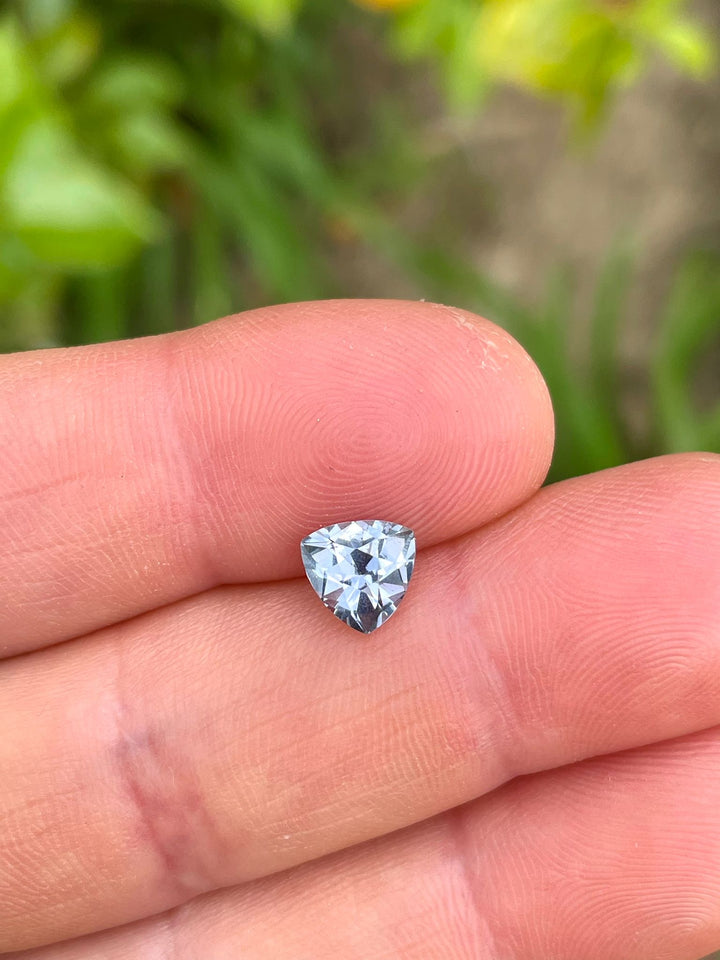 Close-up of a custom trillion shaped sapphire weighing 1.50 carats, showcasing its unique gray, purple, and blue colors, held in fingers with blurred greenery in the background.