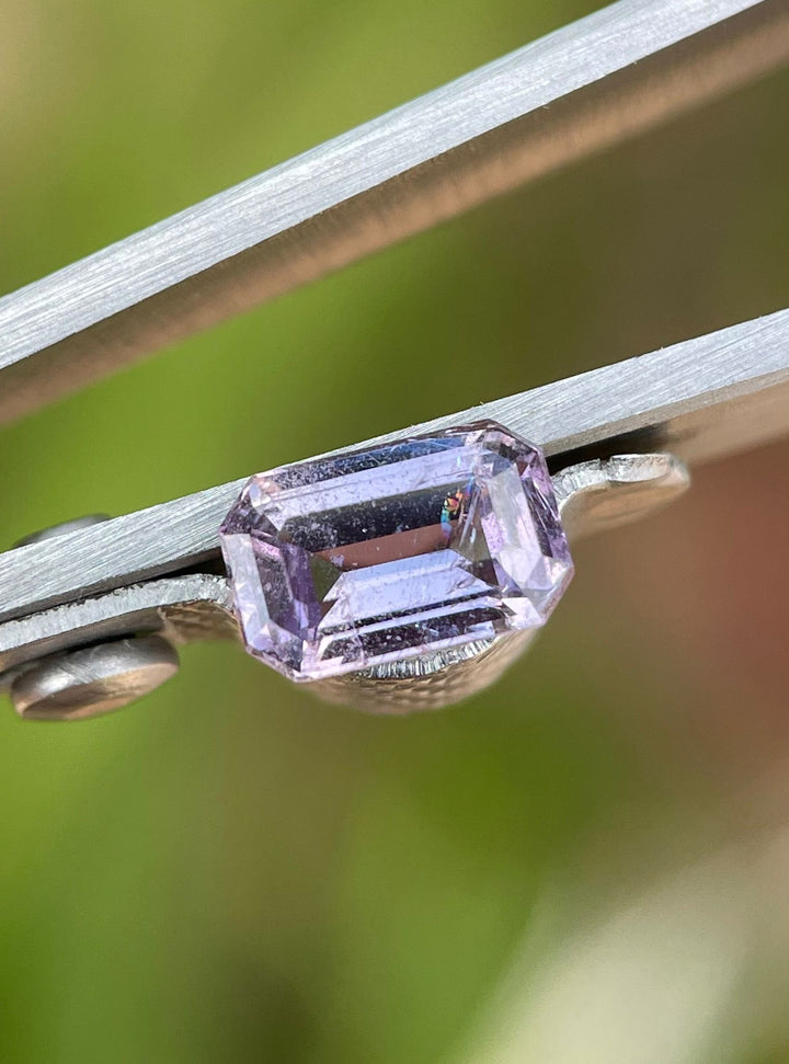 Close-up of a brown and purple sapphire with a modified emerald cut, showcasing its unique facets and clarity, secured in tweezers against a blurred green background.