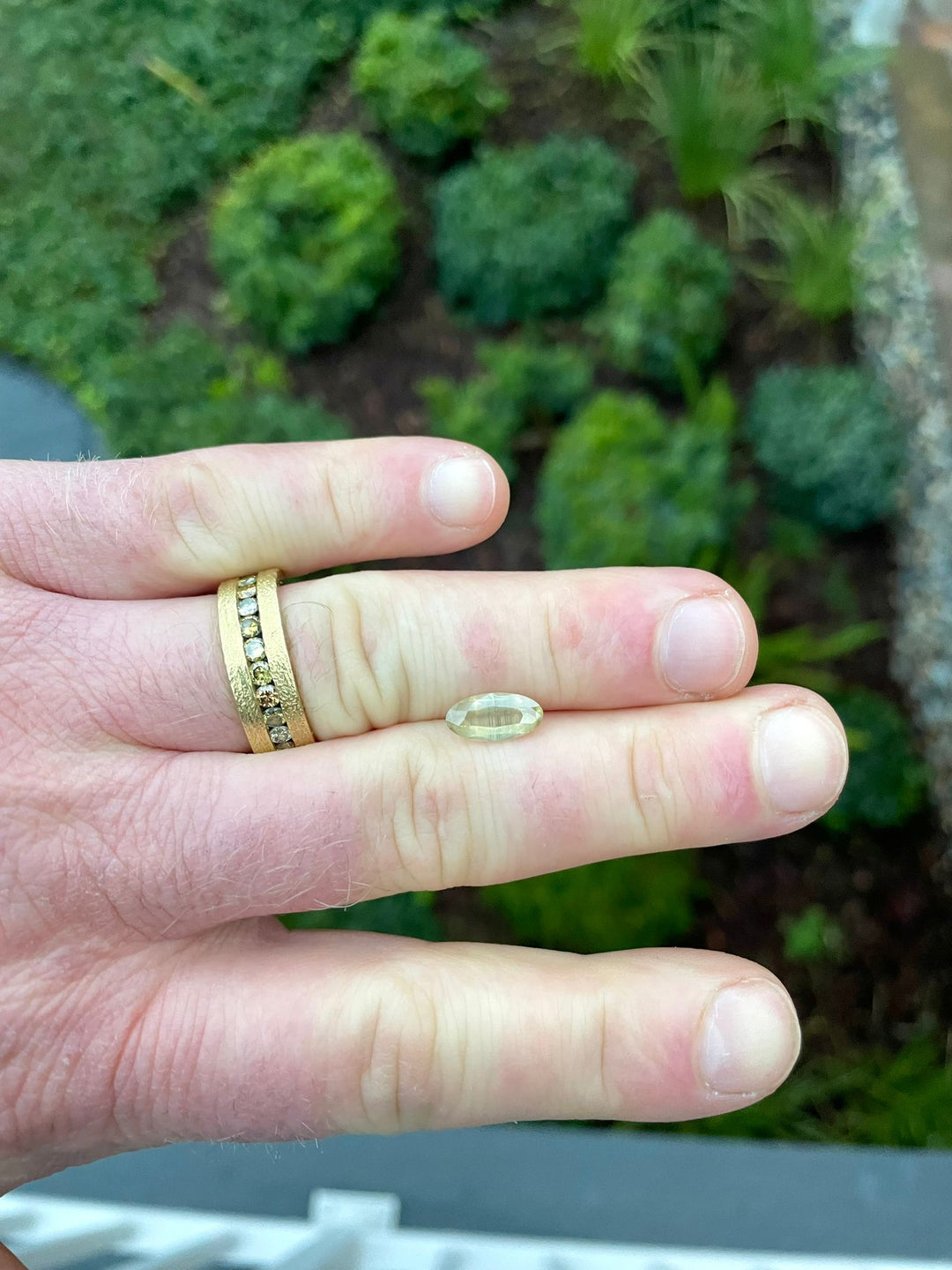Hand holding a 1.93 carat peach, opalescent sapphire with a custom elongated oval cut against a green garden backdrop, showcasing the gem's delicate hue and elegant shape.