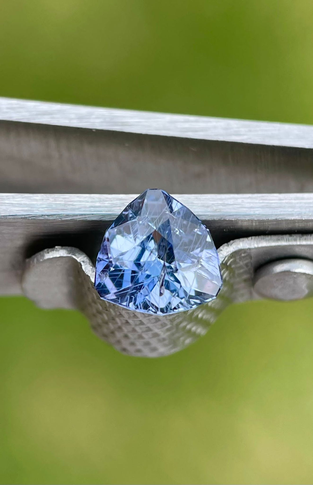 Close-up of a 1.38 carat violet and denim sapphire, showcasing the gemstone's sharp angles and intricate cuts, held securely in tweezers against a green blurred background.