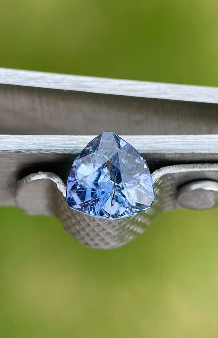 Close-up of a 1.38 carat violet and denim sapphire, showcasing the gemstone's sharp angles and intricate cuts, held securely in tweezers against a green blurred background.