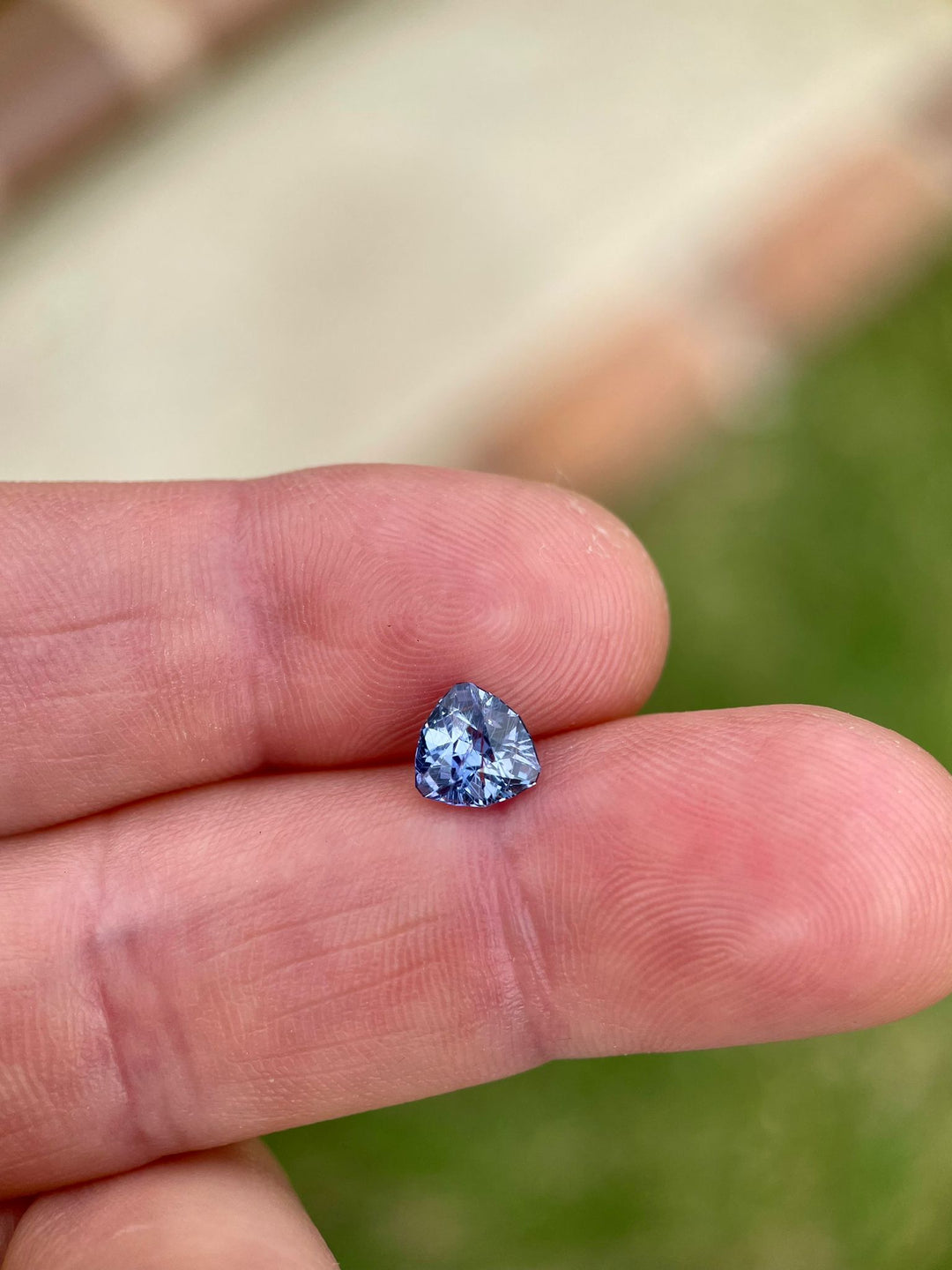 Vivid violet and denim sapphire held between fingers, displaying its unique trillion shape and brilliant color against a soft green backdrop.