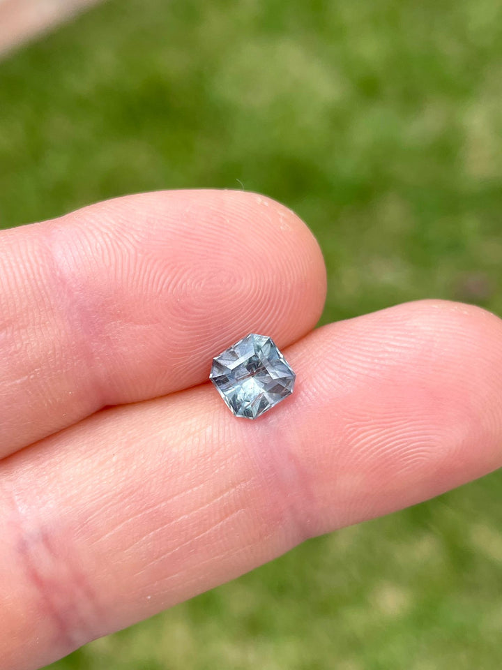 Teal, blue, and gray 1.54 carat modified Asscher cut sapphire, displayed in hand against a grassy backdrop, highlighting its distinct geometric facets and clarity