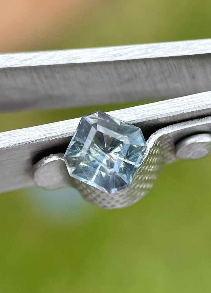 Close-up of a 1.54 carat sapphire with a modified Asscher cut, held securely in tweezers with a green blurred background, showcasing its unique color blend.