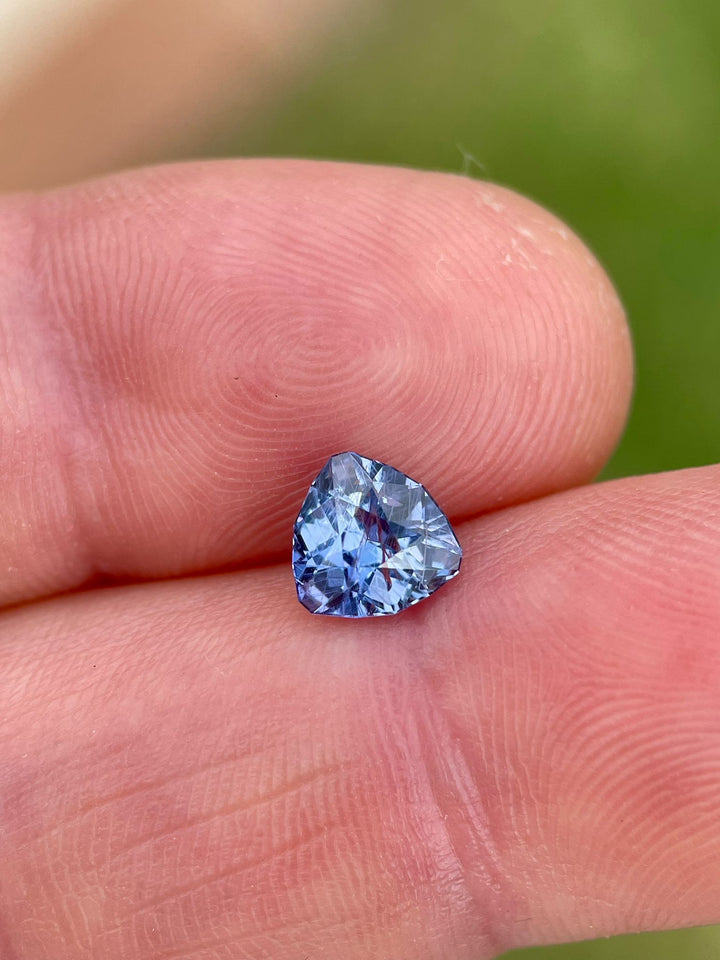 Vivid violet and denim sapphire held between fingers, displaying its unique trillion shape and brilliant color against a soft green backdrop.