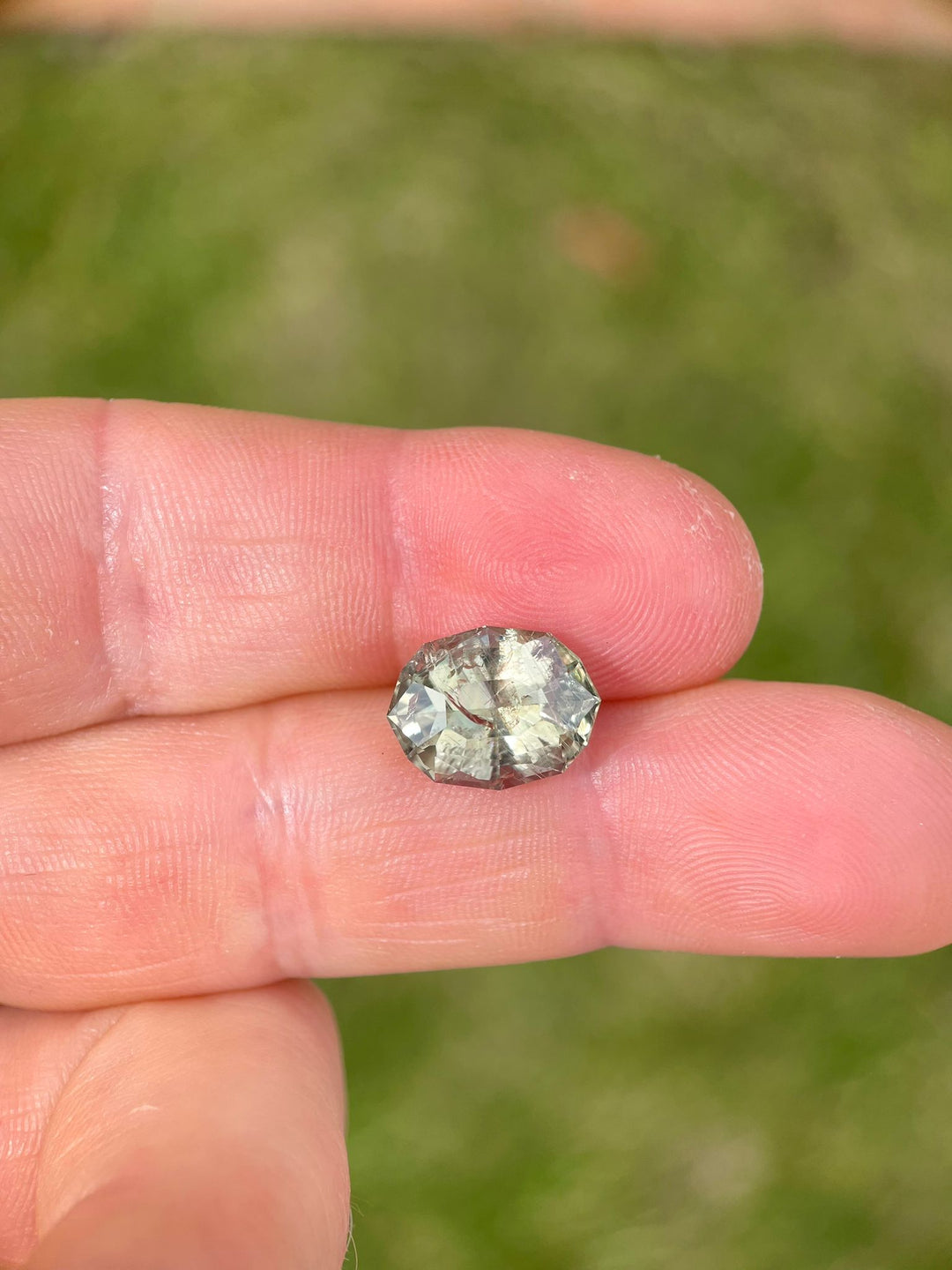 Close-up of a color-changing sapphire held in hand, showcasing the gemstone's natural, untreated appearance and its striking custom geometric oval cut, reflecting light and color variations.
