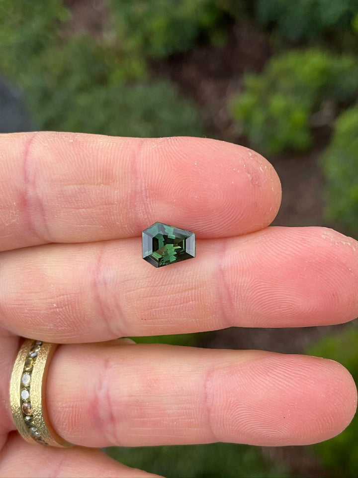 Hand-held view of a 2.38-carat teal green sapphire, illustrating its compact size, unique coffin shape, and the intense color depth, set against a natural outdoor setting