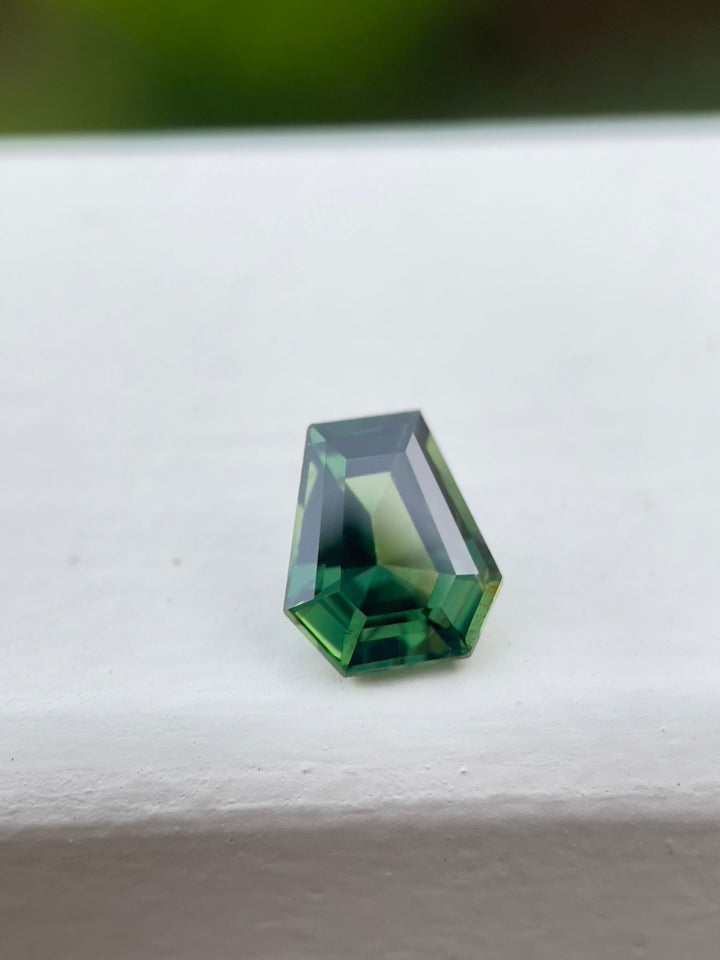 Teal green sapphire resting on a white surface, demonstrating its clear and polished step cut coffin shape, highlighting the gemstone's natural beauty and unheated treatment from Madagascar.