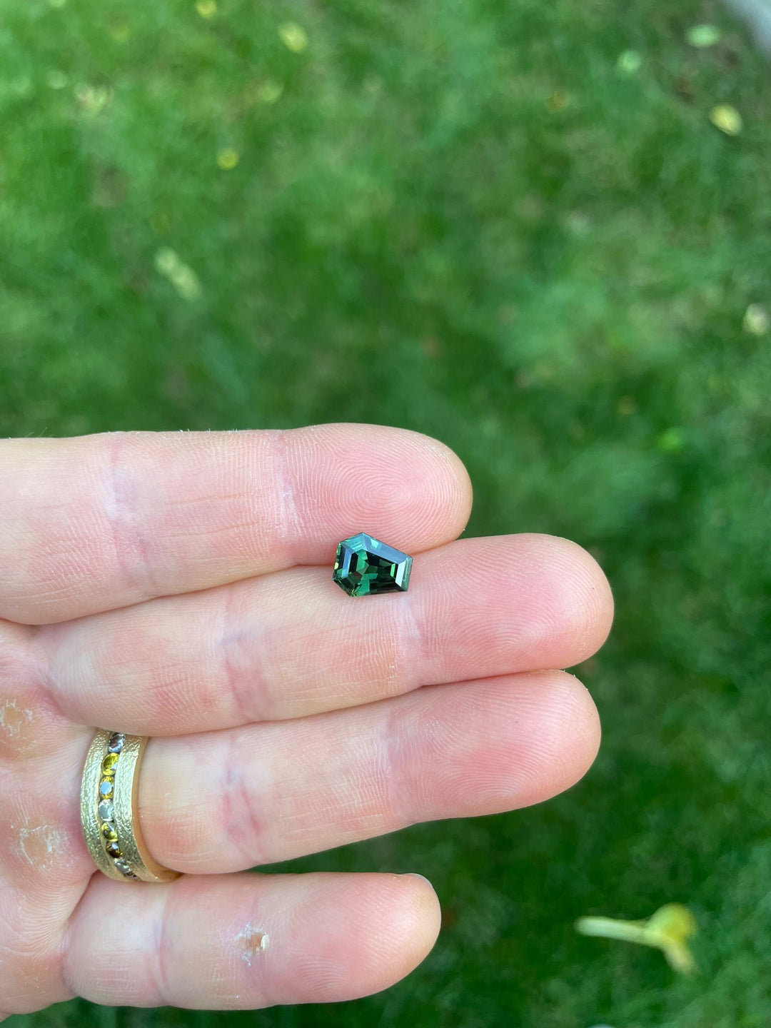 Close-up of a teal green sapphire held between fingers, illustrating the gemstone's unique step cut coffin shape and deep, rich color, against a natural green backdrop.
