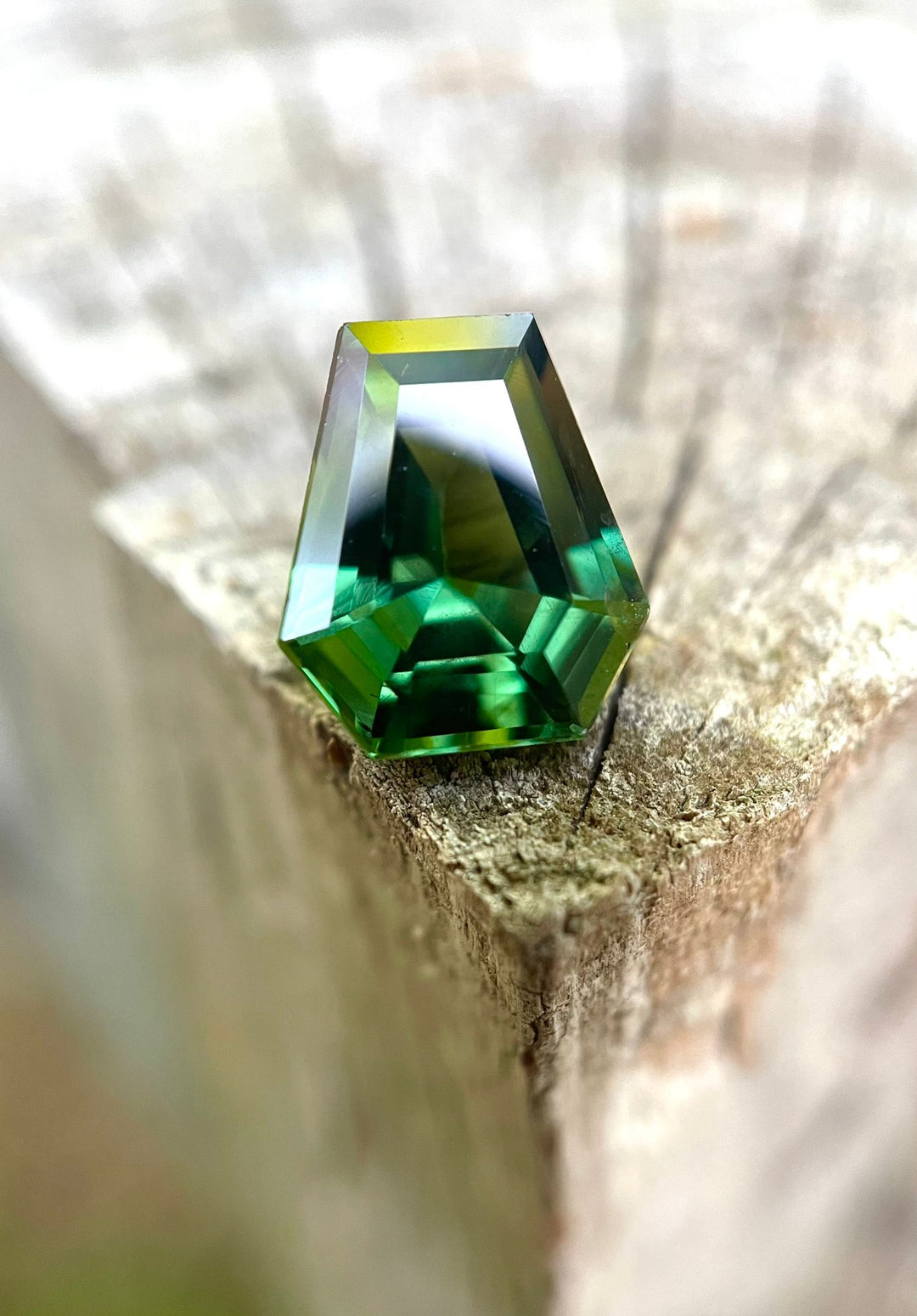 A unique 2.38-carat step cut coffin-shaped sapphire, showcasing vibrant teal and green hues, elegantly perched on a rustic wooden edge with soft blurred background.