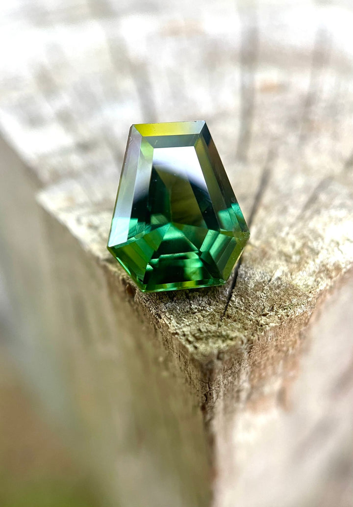 A unique 2.38-carat step cut coffin-shaped sapphire, showcasing vibrant teal and green hues, elegantly perched on a rustic wooden edge with soft blurred background.