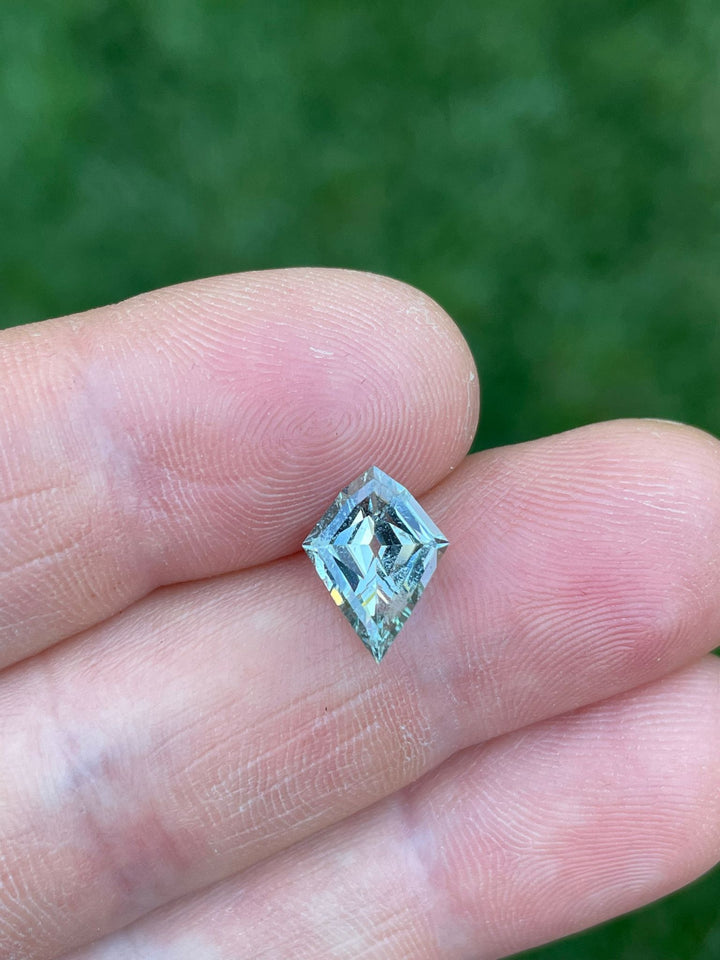 Hand-held view of a green seafoam sapphire, emphasizing the gemstone's geometric modified step cut kite shape and its deep, transparent green color, demonstrating its high quality and natural, untreated state.