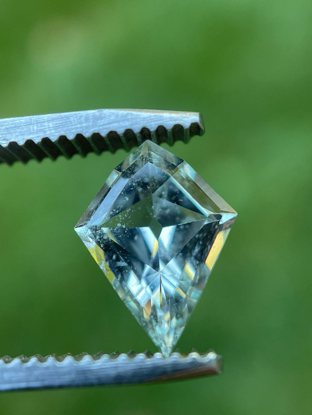 Close-up view of a 2.28-carat sapphire held in tweezers, featuring a geometric modified step cut kite shape with vibrant green and seafoam colors, sourced from Sri Lanka, set against a blurred green background.