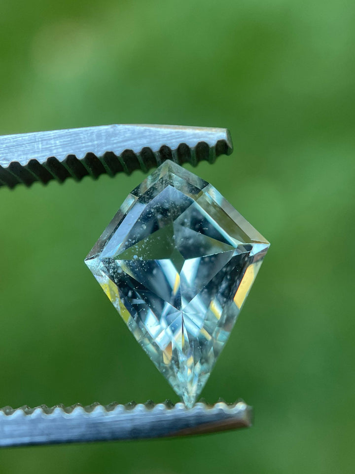 Close-up view of a 2.28-carat sapphire held in tweezers, featuring a geometric modified step cut kite shape with vibrant green and seafoam colors, sourced from Sri Lanka, set against a blurred green background.