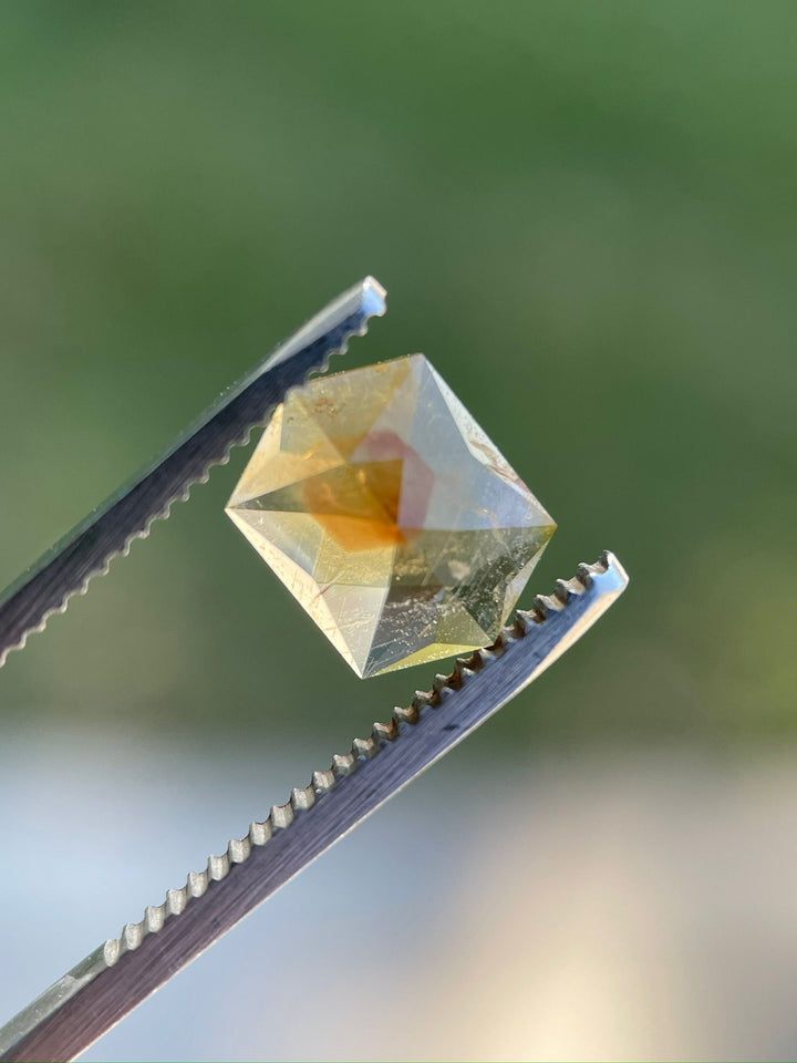 Close-up of a 3.12-carat amber, orange, mauve, and brown sapphire held by tweezers, featuring a custom hexagon rose cut. This gemstone from the Umba River, Tanzania, displays unique color variations and clarity against a blurred green background.
