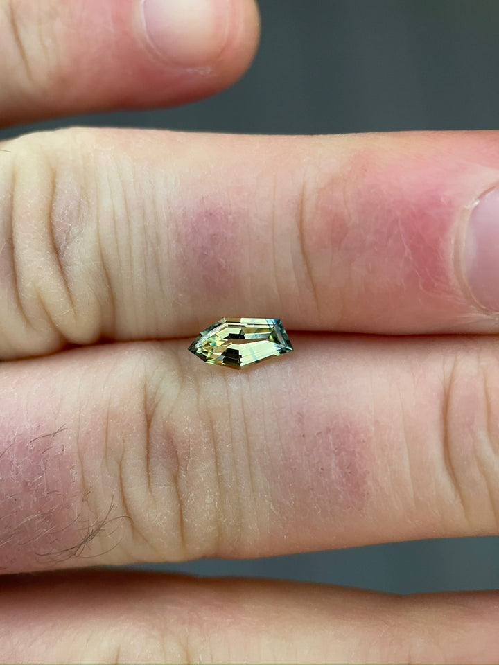 Close-up of a 0.80-carat yellow-blue sapphire resting on a human hand, highlighting the gem's freeform geometric shape and natural, untreated appearance.