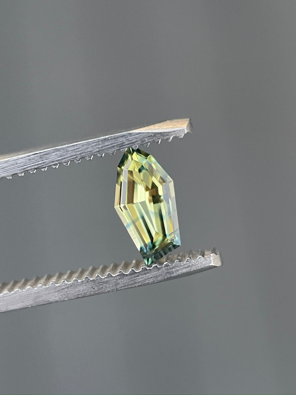 Detailed view of a 0.80-carat sapphire, held by tweezers, displaying a unique freeform geometric cut with striking yellow and blue colors, sourced from Gombe, Nigeria.