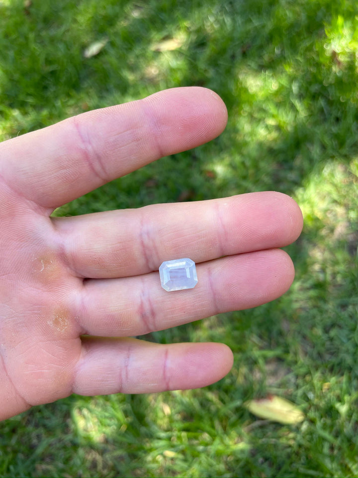 Close-up view of a 5.10-carat opalescent sapphire held in a person's fingers, demonstrating the gem's octagonal step cut and subtle periwinkle tones against a natural green background.