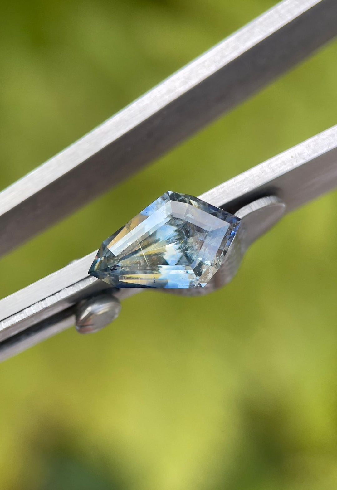 Detailed view of a 2.32-carat geometric step cut kite sapphire, delicately held in tweezers, showcasing its unique colors and precision cut, set against an outdoor setting.