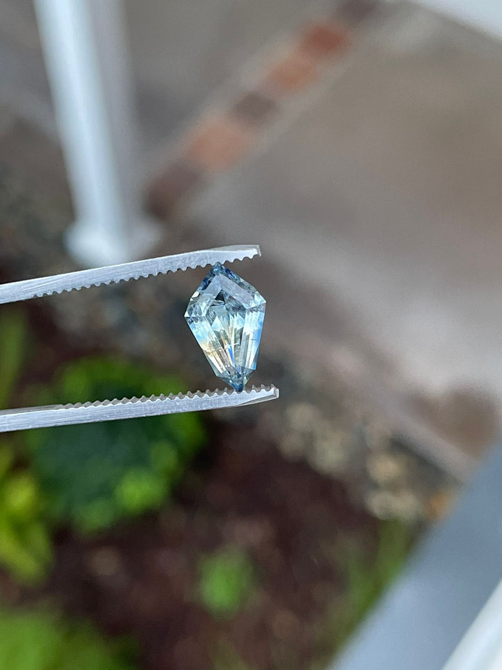 Captivating 2.32-carat geometric step cut kite sapphire held in tweezers, featuring a unique blend of yellow and Ceylon denim blue colors, against a soft green background.