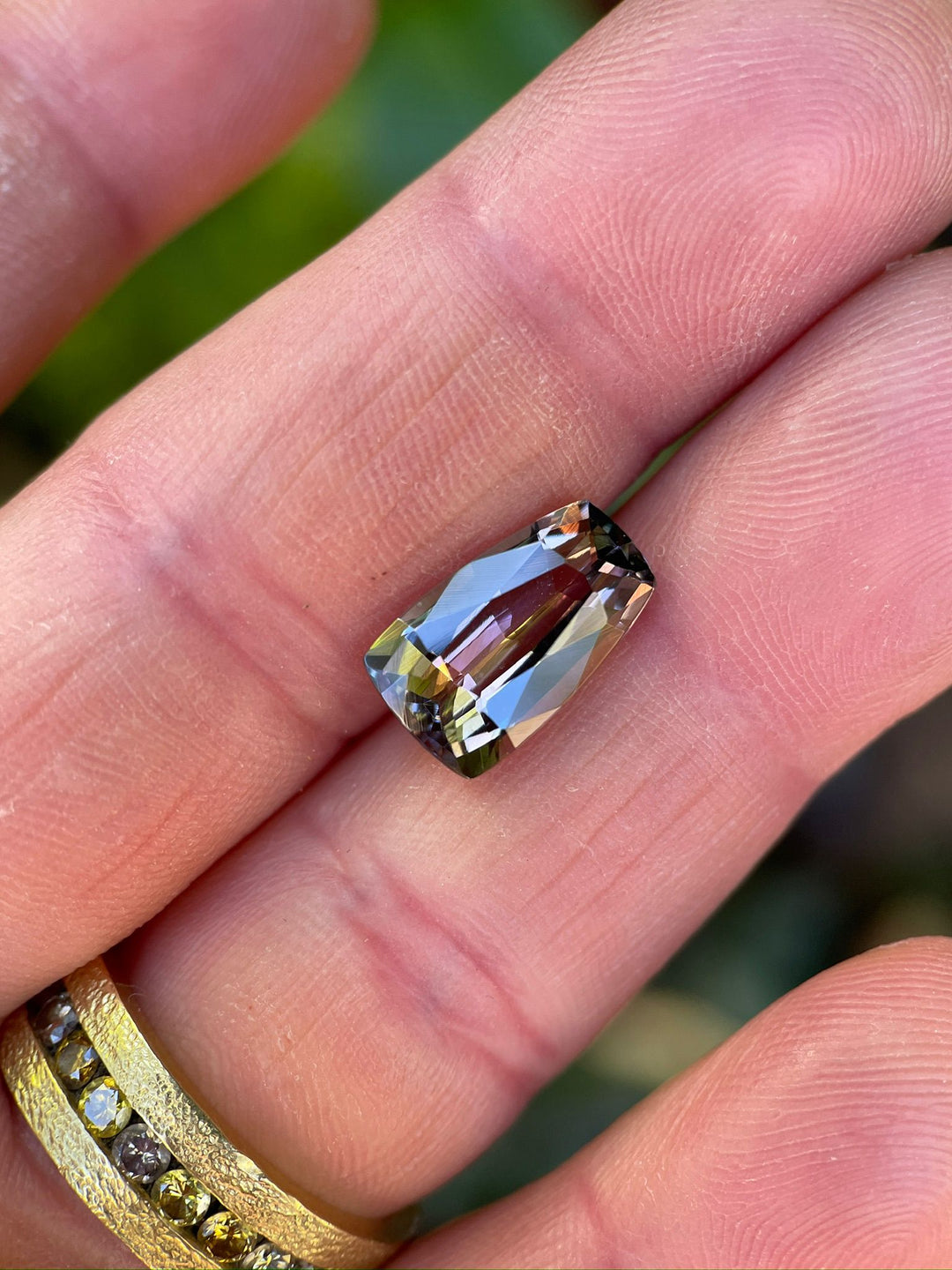 Close-up of a Tanzanite gemstone held in a person’s hand, featuring a custom tapered shield mix cut, weighing 4.98 carats, displaying vibrant color transitions from purple to champagne.