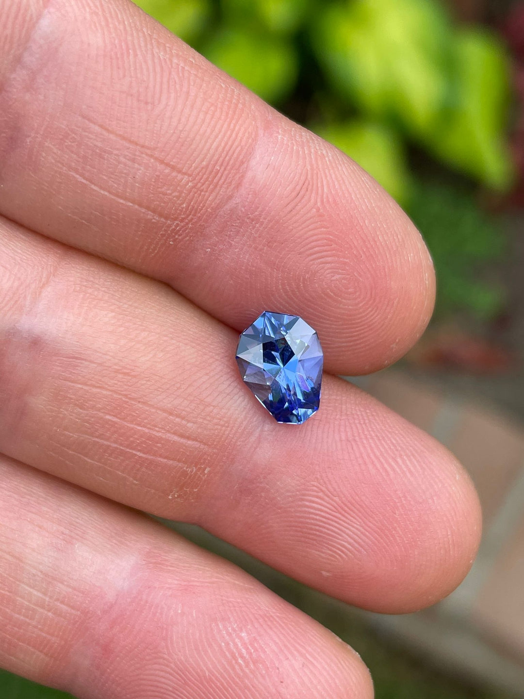 Lush green backdrop with a 2.26-carat ultra violet blue Tanzanite gemstone in a custom geometric teardrop shape held between fingertips, highlighting its vivid color and brilliance, sourced from the Merelani Hills in Tanzania.