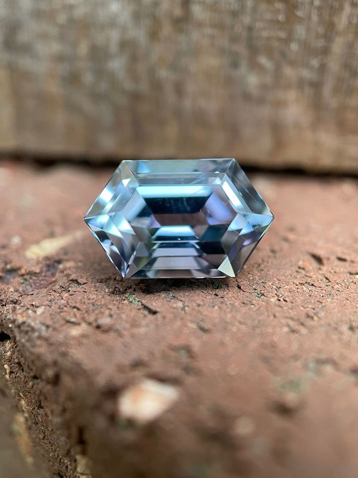Tanzanite gemstone resting on a textured brick surface, showcasing its unique step cut hexagonal shape and mesmerizing color transition from teal to purple to blue. Weighing 1.79 carats and sourced from Merelani Hills, Tanzania, this gem measures 9.54 x 6.38 x 4.38 mm and is valued at $1700.50.