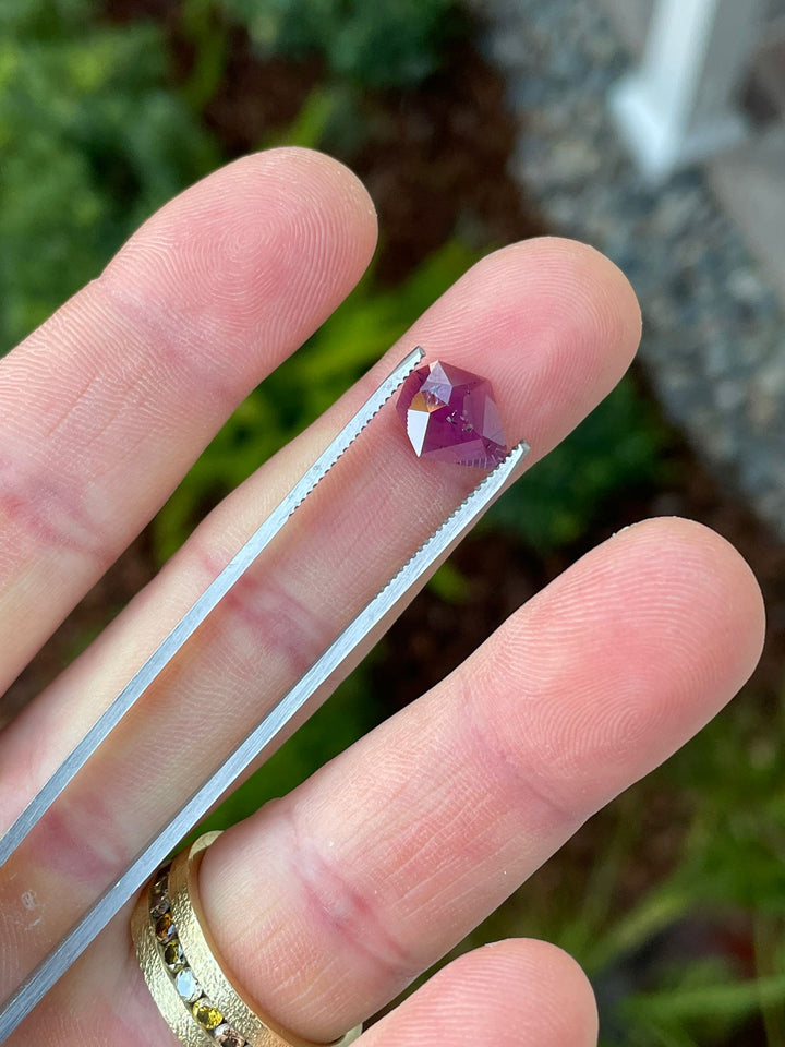 Close-up of a 2.38-carat mulberry purple sapphire held in tweezers over a garden background, featuring a custom shield rose cut, sourced from the Umba River, Tanzania, priced at $1,547.